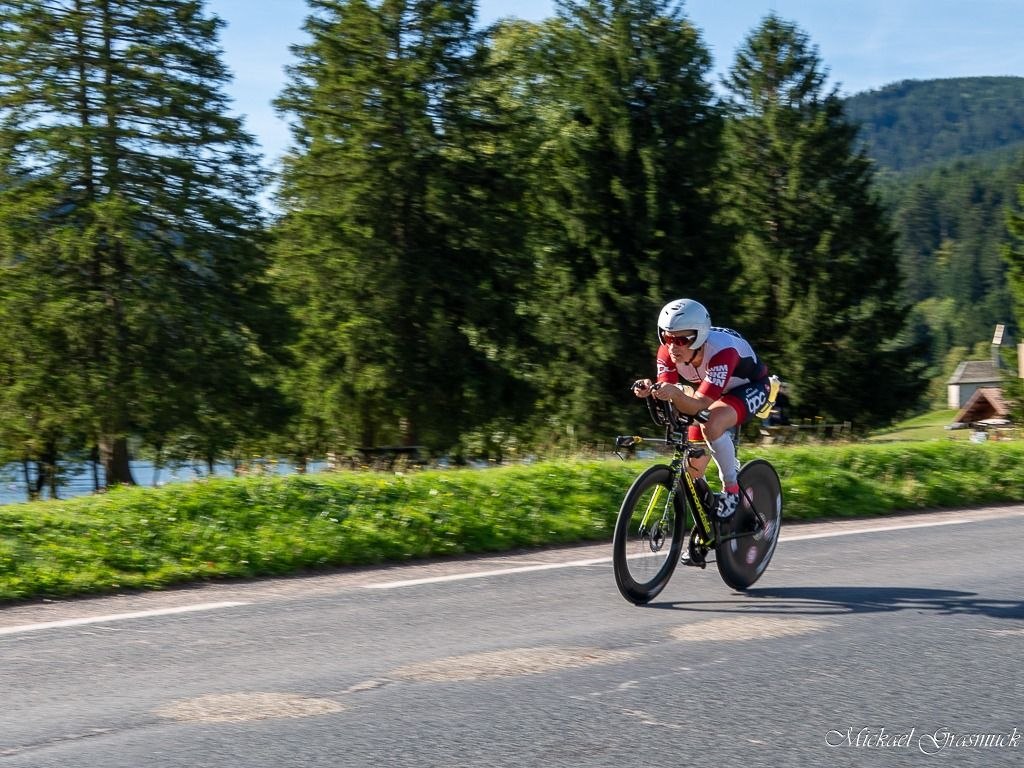Triathlète BPC en position aérodynamique sur une route bordée de forêt et de relief vosgien.